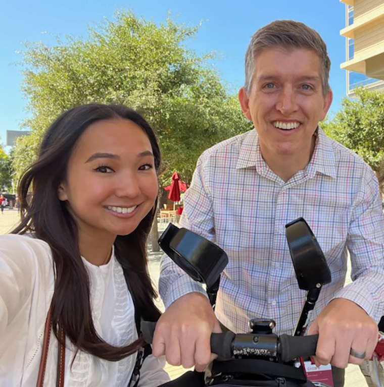 A young woman poses next to a middle-aged man in a mobility assistance device.