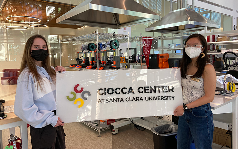 Two masked young women hold up a plastic sign for the Ciocca Center.