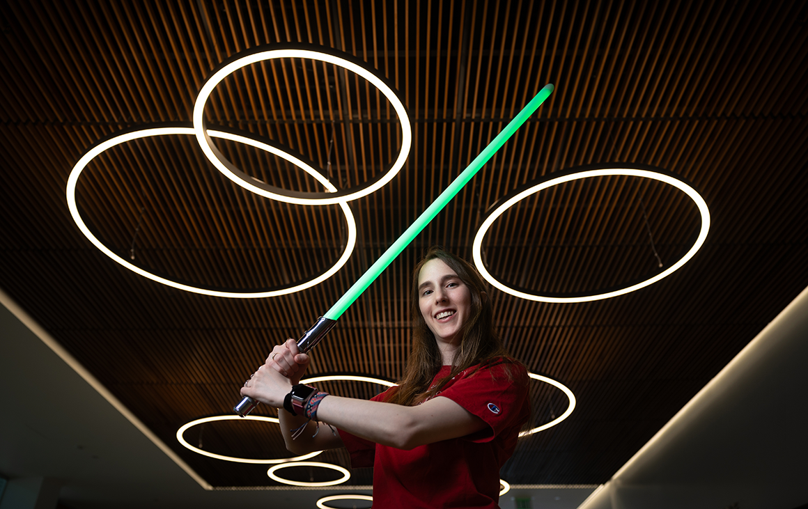 A woman wields a glowing green lightsaber against a striped wooden ceiling with circular LED lights.