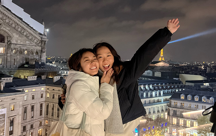 Two young women pose closely in front of the Paris cityscape at night.